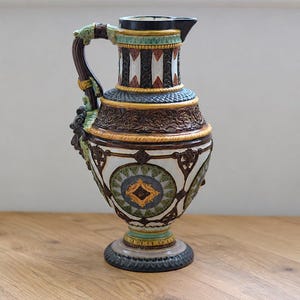 Könnte beinhalten: Ein Keramikkrug mit einem dunkelbraunen Henkel und einem mehrfarbigen Körper mit komplexen Reliefmustern. Das Design umfasst weiße, grüne, blaue und goldene Akzente mit geometrischen Formen und floralen Motiven auf einem dunklen Sockel.