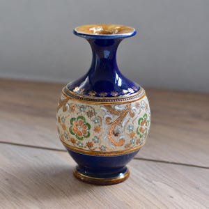 May include: A ceramic vase featuring a deep cobalt blue neck and base with a central band of cream-coloured textured material. The central section is decorated with intricate floral patterns in metallic gold, green, and white, accented by small gold stars.
