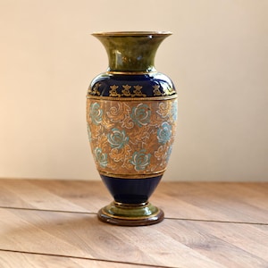 May include: A ceramic vase featuring a flared rim and a tapered base in mottled olive green. The body is deep blue with a central band of gold filigree and light blue floral patterns, accented by thin gold horizontal lines and decorative motifs.