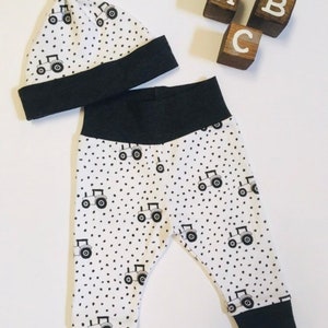 May include: A white and black baby outfit with a tractor print. The outfit includes a hat and pants. The hat has a black trim and the pants have black cuffs.