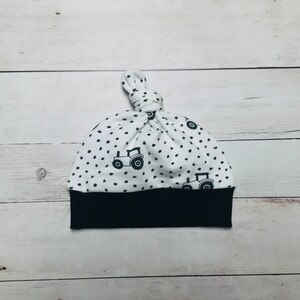 May include: White baby hat with a black and white tractor print and black polka dots. The hat has a black band at the bottom and a knot at the top.