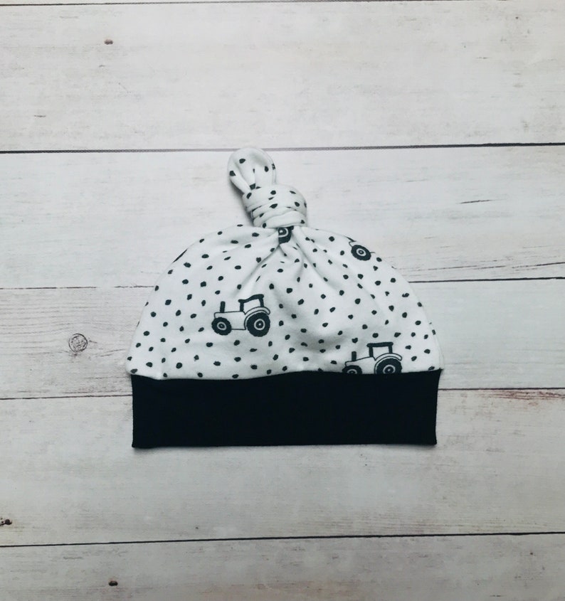 May include: White baby hat with a black and white tractor print and black polka dots. The hat has a black band at the bottom and a knot at the top.
