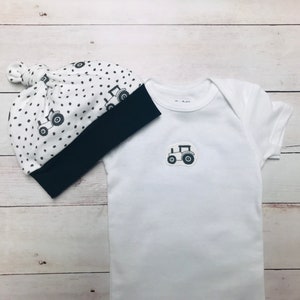May include: A white baby onesie with a black tractor design and a white and black polka dot hat with a navy blue brim.