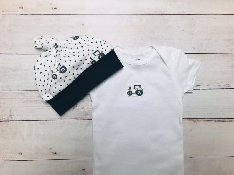 May include: A white baby onesie with a black tractor design and a white and black polka dot hat with a navy blue brim.
