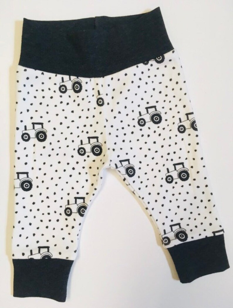 May include: White baby leggings with a black tractor and polka dot pattern. The leggings have black elastic at the waist and ankles.