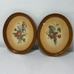 May include: A pair of oval wall frames with brown wooden borders, each containing a botanical print of a floral bouquet on a cream-colored background. The prints feature detailed illustrations of red roses, green leaves, and small blue flowers.