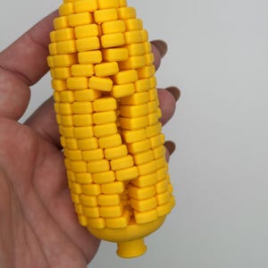May include: A bright yellow, three-dimensional corn cob figure featuring a textured surface composed of individual rectangular kernels arranged in vertical rows, with a small, rounded protrusion at the top and a cylindrical base at the bottom.