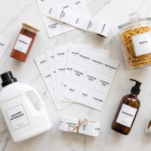 Peut inclure: Une collection d'étiquettes adhésives blanches avec du texte noir incluant des mots comme Pasta, Shampoo et Laundry Detergent, présentées avec un bocal en verre, un pot à épices, un bidon de lessive blanc et un flacon pompe ambré étiqueté.