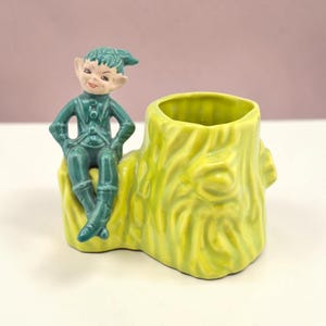 May include: A ceramic planter shaped like a yellow tree stump with a dark green elf figure sitting on the side. The elf has pointed ears, a cap, and a smooth glazed finish. The tree stump features a textured, bark-like surface and a hollow center.