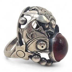 May include: A silver ring featuring an intricate openwork design of two stylised fish figures. The metalwork includes engraved scales and circular accents. A large, smooth, deep red cabochon gemstone is set prominently at the front of the ring.