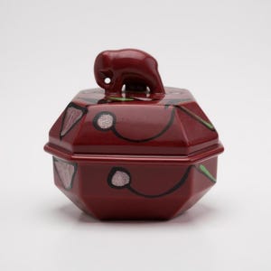Peut inclure: Un pot en céramique octogonal rouge foncé avec un couvercle brillant. Le couvercle est surmonté d'une figurine d'éléphant sculptée. Le corps et le couvercle sont ornés de dessins au trait noir représentant des cerises et des feuilles.