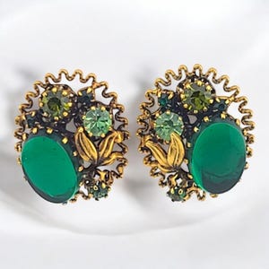 May include: A pair of oval-shaped earrings featuring a dark gold-toned metal frame with a wavy, scalloped edge. Each earring is adorned with a large, smooth, dark green oval cabochon, smaller round light green crystals, and gold metal leaves.