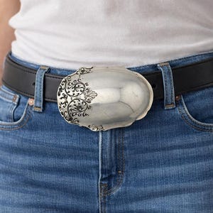Könnte beinhalten: Ein schwarzer Ledergürtel mit einer großen, ovalen Gürtelschnalle aus silberfarbenem Metall. Die Schnalle hat eine glatte, polierte Oberfläche auf der rechten Seite und ein komplexes, erhabenes Rankenmuster auf der linken Seite, befestigt mit einem Metalldorn.
