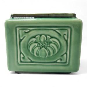 May include: A rectangular ceramic planter with a glossy light green glaze. The front face features a raised relief design of a stylised flower centred within a square border. The top and bottom edges are finished with a dark brown trim.