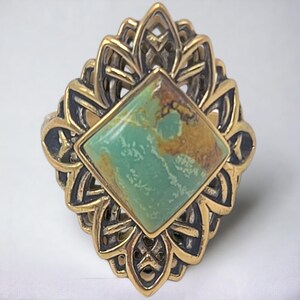 Peut inclure: Une bague avec une pierre turquoise carrée centrale présentant des veines de matrice brune, sertie dans une monture en métal doré ajouré et ornée, avec des détails sombres et en creux sur les bords du motif métallique complexe.