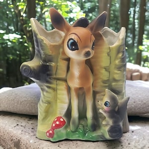 May include: A ceramic planter shaped like a hollow tree stump featuring a tan deer standing in the center with large blue eyes and dark ear tips. A small gray squirrel sits at the base next to a red mushroom with white spots on a green patch.