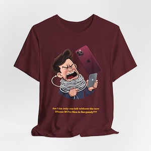 May include: A burgundy short-sleeve t-shirt featuring a cartoon illustration of a person tangled in white charging cables, holding a gray smartphone, with a burgundy smartphone floating above. Text reads: Am I the only one left without the new iPhone 18 Pro Max in Burgundy???