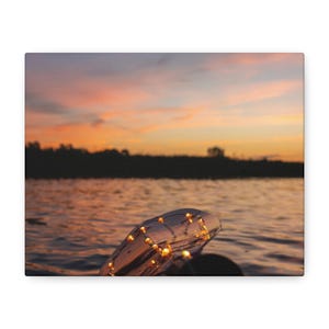 May include: A clear glass bottle containing a string of small, glowing warm-white LED fairy lights. The bottle is positioned against a backdrop of a calm body of water and a vibrant sunset sky featuring shades of orange, pink, and purple hues.
