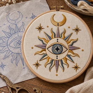 May include: An embroidery hoop containing a fabric panel with a stitched design of a central eye surrounded by sun rays in gold, blue, and purple thread. A crescent moon sits above the eye, with small stars and dots surrounding the central motif.