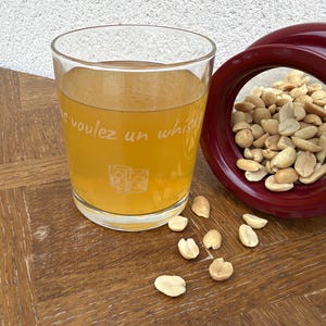 Peut inclure: Un verre transparent rempli d'un liquide ambré, portant l'inscription blanche "voulez un whisky" et un petit graphique de deux verres. À côté se trouve un bol en céramique rouge foncé rempli de cacahuètes, avec quelques arachides éparpillées.