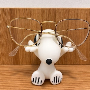 Könnte beinhalten: Eine goldgerahmte Brille mit klaren Gläsern, die auf einer weißen Hunde-Figur im Cartoon-Stil ruht. Die Figur hat schwarze Ohren, eine schwarze Nase und schwarze Pfotenabdrücke auf den Füßen, um die Brille sicher an ihrem Platz zu halten.