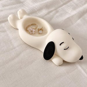 May include: A cream-colored ceramic dog-shaped trinket dish with black ears and nose, featuring a recessed center holding a gold-toned ring with a white flower detail and a small gold-toned stud earring with a clear stone.