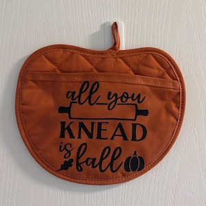 May include: An apple-shaped quilted pot holder in burnt orange fabric with a pocket. The black text reads "all you KNEAD is fall" with a rolling pin graphic, an oak leaf, and a small pumpkin icon. It features a fabric loop for hanging.