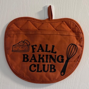 May include: An apple-shaped, quilted orange oven glove featuring a black graphic of a slice of pie and a whisk. The text "FALL BAKING CLUB" is printed in black lettering across the centre. The glove includes a fabric loop for hanging.