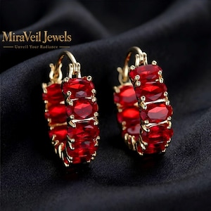 May include: A pair of gold hoop earrings featuring a row of oval-cut vibrant red gemstones set in a prong setting. The text MiraVeil Jewels Unveil Your Radiance is visible in the upper left corner of the image.