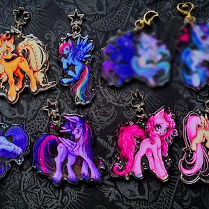 May include: A collection of eight acrylic keychains featuring colorful pony illustrations with various poses and expressions. Each charm includes a metal clasp, with shapes including stars and flowers, showcasing vibrant characters in shades of orange, blue, purple, and pink.