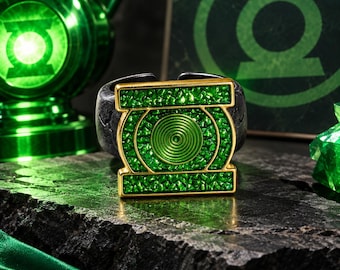 Puede incluir: Un anillo que presenta un emblema cuadrado con un borde dorado y un centro verde brillante texturizado. El emblema muestra un símbolo circular verde con anillos concéntricos. La banda del anillo es oscura, mate y texturizada.
