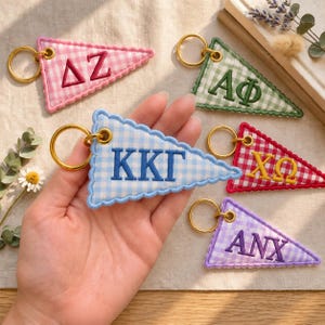 May include: Five triangular pennant keychains with gold-tone metal rings. Each features a gingham check pattern and embroidered Greek letters: pink with ΔΖ, green with ΑΦ, blue with ΚΚΓ, red with ΧΩ, and purple with ΑΝΧ. All have scalloped edges.