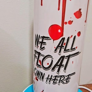 May include: A white cylindrical tumbler featuring a red balloon, red blood-like drips, and a paper boat graphic. The text "WE ALL FLOAT DOWN HERE" is printed in black, stylized lettering. The tumbler has a clear lid and a smooth, glossy finish.