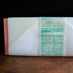 May include: A roll of white Pellon SK135 Sheer-Knit specialty apparel fusible interfacing. The product is wrapped in a paper band featuring green text and instructions, with a portion of the translucent white fabric extending from the roll.