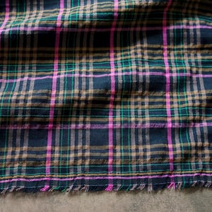 May include: A woven fabric featuring a dark navy blue base with a plaid pattern consisting of intersecting horizontal and vertical lines in bright pink, forest green, and tan. The bottom edge of the textile shows a raw, frayed hemline.