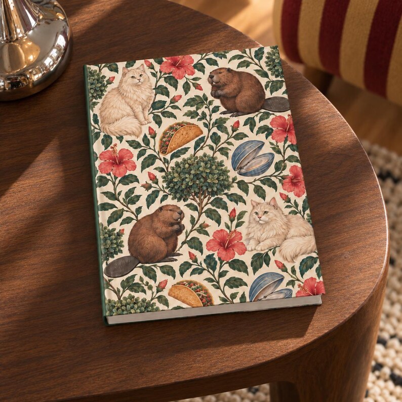 Styled digital mockup of the botanical hardcover journal on a curved wooden side table, beside a chrome lamp and a striped chair. Cream cover with green foliage, pink flowers and animal and food motifs. Furniture and props are not included.