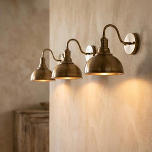 Pode incluir: Um candeeiro de parede triplo com três abajures em forma de cúpula com acabamento em latão antigo. Cada abajur está ligado a um suporte de parede circular por um braço curvo, emitindo uma luz quente direcionada para baixo.