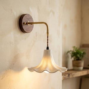 Könnte beinhalten: Eine Wandleuchte mit einem runden Sockel aus dunklem Holz, einem messingfarbenen Metallarm und einem gedrehten braunen Kabel. Die Leuchte besitzt einen weißen, geriffelten, glockenförmigen Keramikschirm, der ein warmes Licht ausstrahlt.