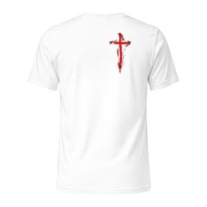 May include: A white short-sleeved t-shirt featuring a red cross graphic on the upper left chest area. The cross design has a distressed, brushstroke appearance with vertical and horizontal lines that taper into textured, paint-like edges.