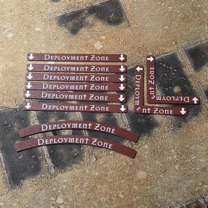 May include: A collection of brown game markers with white text reading Deployment Zone. The set includes six straight rectangular markers, two L-shaped corner markers, and two curved markers, all featuring white arrows indicating direction.