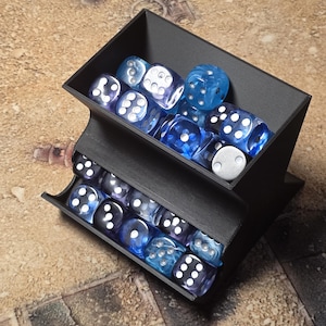 May include: A black plastic two-tiered dice storage rack filled with various translucent blue, purple, and clear six-sided dice. The rack features an open top bin and a lower shelf, both containing multiple dice with white pips, organized in a compact display.