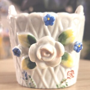 May include: A white ceramic egg cup featuring a raised lattice texture, adorned with a central sculpted white rose, green leaves, and small blue flowers. The base includes a small red floral stamp and delicate painted green and blue accents.
