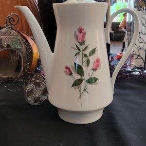 May include: A white porcelain coffee pot featuring a delicate floral design of three pink rosebuds with green stems and leaves. The pot has a tall, tapered body, a curved handle, a long spout, and a matching lid with a small round knob on top.