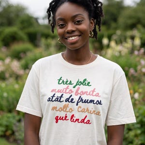 May include: A cream-colored short-sleeve t-shirt featuring five lines of embroidered text in different colors. The text reads: très jolie in green, muito bonita in pink, atât de frumos in dark blue, molto carina in tan, and qué linda in red.