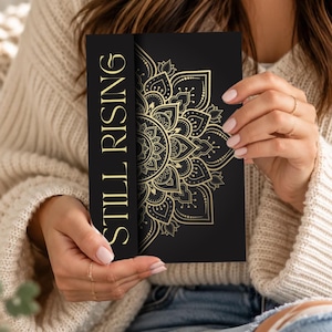 May include: A rectangular black card featuring the gold text STILL RISING vertically on the left side and a detailed gold mandala pattern on the right side. The design is printed on a smooth surface with high-contrast metallic gold ink.