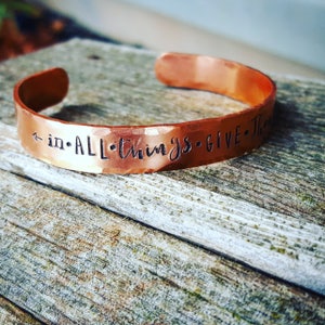 Puede incluir: Una pulsera de cobre con las palabras "in all things give thanks" grabadas.