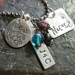 May include: A silver necklace with multiple charms, including a round charm that says "Keep Calm & Clog On", a rectangular charm that says "J4C", a rectangular charm that says "Nicole", and a blue glass bead.
