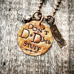 May include: A copper necklace with a charm that says "Don't Do Dumb Stuff" and a smaller charm that says "Mom" with a heart. The necklace is on a wooden background.