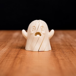 May include: A cream-coloured, three-dimensional ghost figurine featuring a segmented, layered design. The figure has two small arms extended outwards, a rounded head, and a carved facial expression with wide, sad eyes and an open mouth.