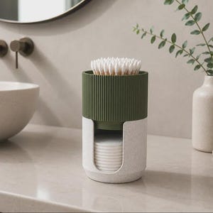 Puede incluir: Un organizador de baño cilíndrico con una sección superior acanalada en verde oliva oscuro para bastoncillos y una base de color blanco roto moteado con un corte vertical para dispensar discos de algodón redondos.